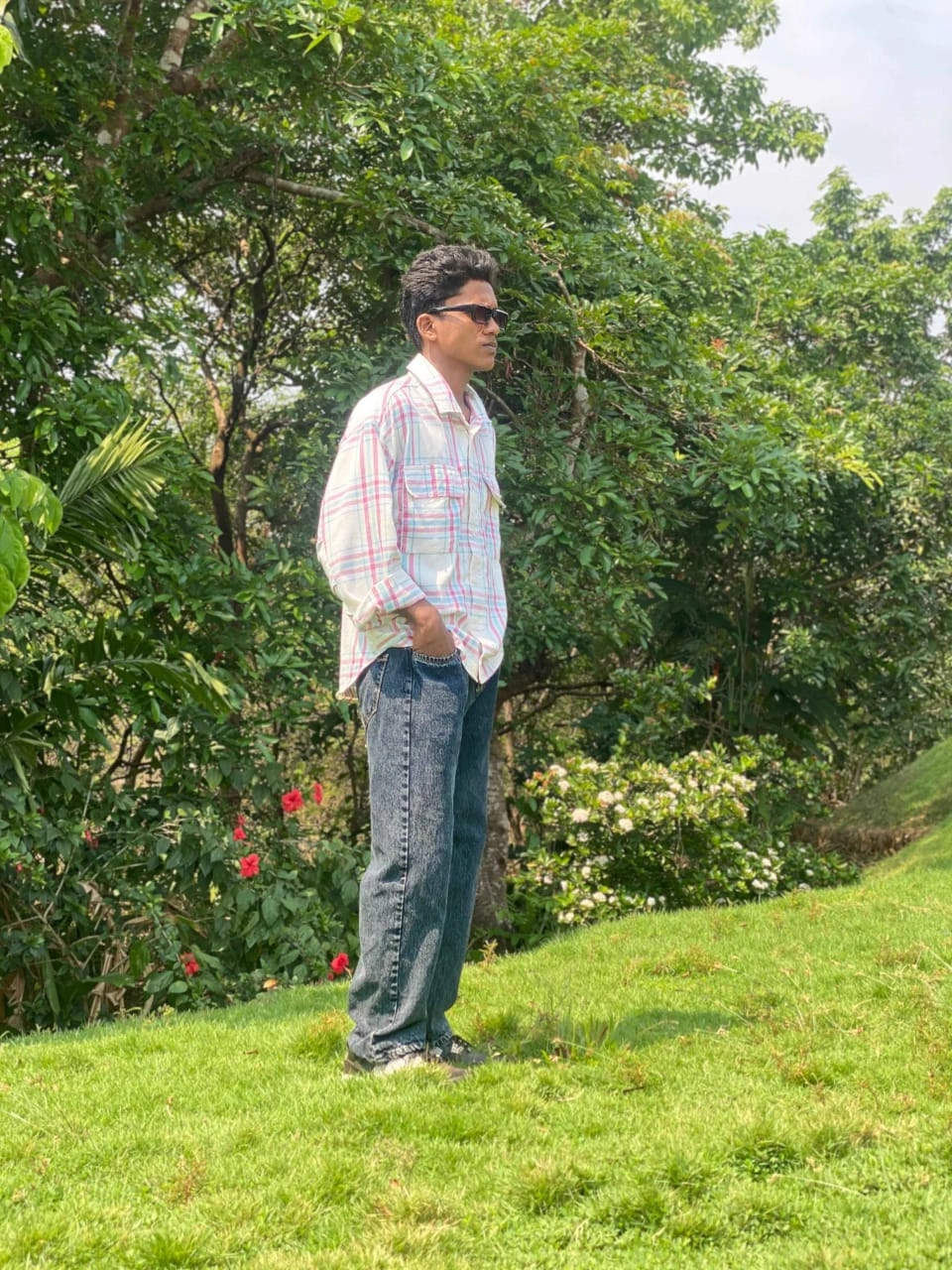 A young man wearing a plaid button-up shirt, jeans, and sunglasses standing outdoors in a lush green garden setting.